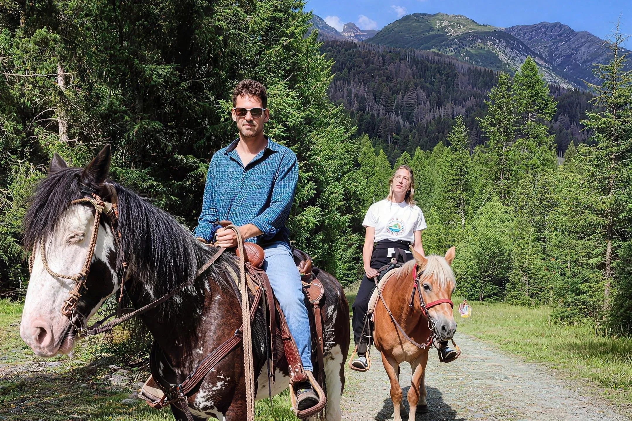 Due cavalieri al passo su una strada sterrata fra gli abeti, con le montagne sullo sfondo